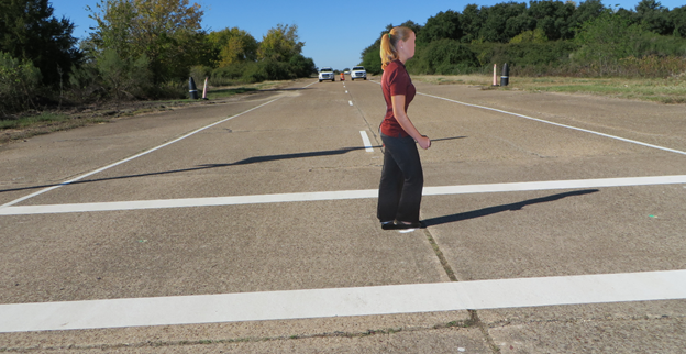 Figure 6. Photo. View of 54-inch cutout pedestrian used in study. This photo shows a view of the 54-inch photograph cutout of a pedestrian in the crosswalk in the middle of the two-lane street at the study site. The cutout is facing the right side. In the background 200 ft away, two vehicles are visible (one per each lane) with an orange work zone barrel between them.
