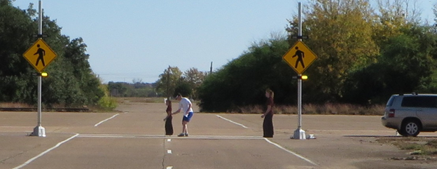 Figure 7. Photo. Researcher removing short cutout pedestrian after placing tall cutout pedestrian. This photo shows a researcher removing the 54-inch photograph cutout of a pedestrian from the center of a crosswalk along a two-lane road at the study site. Two beacons flashing below the pedestrian signs are visible at both sides of the crosswalk, and a 70-inch photograph cutout of a pedestrian is on the right side of the street facing left.