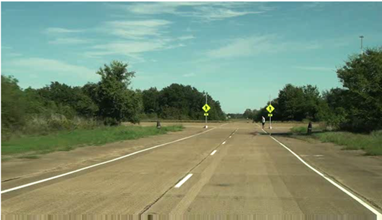 Figure 9. Photo. View at start of the driving video for the concluding survey for queries 1 and 2. This photo shows an image capture of video taken from a vehicle at the closed-course study site from the point of view of an observer 200 ft from the crosswalk. The crosswalk spans a two-lane road and has two signs with rectangular rapid flashing beacons (RRFBs)â€”one on each side of the crosswalk. A researcher is standing on the right side of the road at the crosswalk facing left, and the RRFBs are not activated.