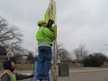 Figure 50. Photo. Installation of the light bar in field. This photo shows a researcher on a ladder installing a set of rectangular rapid flashing beacons below an S1-1 school crossing sign and above a W16-7 left arrow sign.
