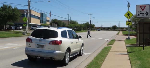 Figure 51. Photo. CS-02 study site with installed temporary light bars and staged pedestrian crossing. This photo shows a pedestrian crossing a road with two lanes for each direction of travel plus a center two-way left-turn lane. In the foreground, there is a car in the far right lane of traffic approaching the crosswalk and yielding to the pedestrian; the yielding position of the car is at the advance yield markings on the pavement upstream of the crosswalk. The crosswalk is marked with a continental marking design. The crosswalk has a R1-5 YIELD HERE TO PEDESTRIAN sign on the right side of the road adjacent to the advance yield markings, and there is a rectangular rapid-flashing beacon assembly on each side of the road at the crosswalk. The assembly consists of a W11-2 pedestrian warning sign, a W16-7 arrow sign, and rectangular rapid flashing beacons located between the W11-2 and W16-7 signs.