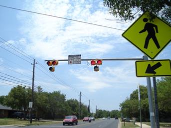 Figure 54. Photo. Example of PHBs being used in Austin, TX. This photo shows pedestrian hybrid beacons (PHBs) at a crosswalk in Austin, TX. Two PHBs are mounted on a mast arm. The faces of the PHBs are two red signal indications side by side with a yellow signal indication centered below them. The signals do not have a back plate. Between the PHBs there is a STOP ON RED sign. In the foreground upstream of the mast arm and pole there is a W11-2 pedestrian crossing sign with a W16-7 arrow sign pointing to the left.