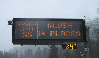 This photo shows a WSDOT DMS on I-90 at Snoqualmie Pass. The screen is black with yellow text. The left side of the screen reads, “SPEED LIMIT 55.” There is a yellow vertical line separating that text from the remaining text, which reads, “SLUSH IN PLACES.” Below the sign, there is a smaller DMS displaying the current temperature, which is 34 degrees Fahrenheit.