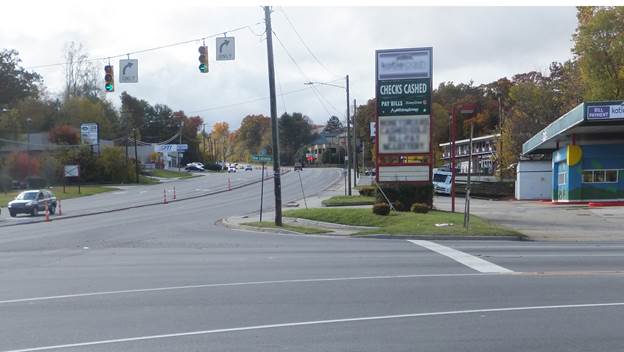 Street level view of two legs of an urban intersection. Immediately after the intersection, there is a driveway entrance to a parking lot on the right. On the left, there is a center median.