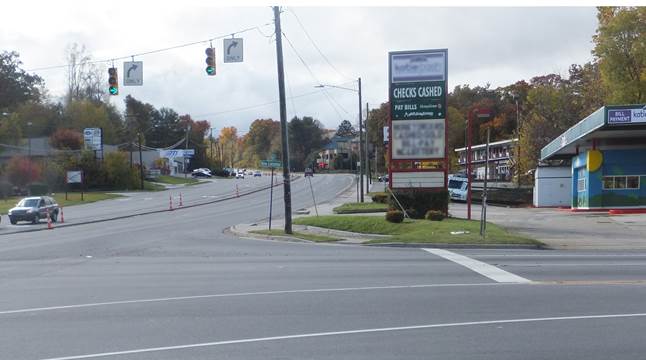 Photo. Signalized intersection with limited receiving corner clearance. Driver’s-eye view of an urban intersection. Immediately after the intersection, there is a driveway entrance to a parking lot on the right. On the left is a center median.
