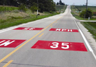 This is a photo of a two-lane road with pavement markings in the middle of each lane that display the speed limit. These pavement markings are boxes of red with the speed limit in white writing.