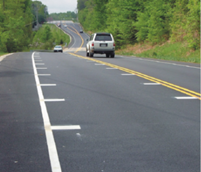 This is a photo of a two-lane road with transverse markings on both sides of the lane (the edge line and the center line). The transverse markings are spaced specifically to give drivers the perception of driving faster.