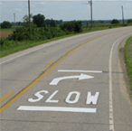 This is a photo of a two-lane road with a pavement marking that reads 'SLOW' with a curved arrow above that indicates there is about to be a curve in the road. The pavement marking has a bar both above the curved arrow and below the wording.