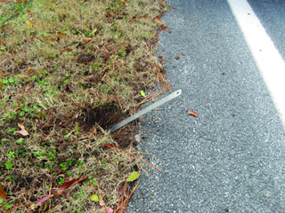 This photograph shows a close-up view of one of the sites that was resurfaced and treated with the safety edge. The edge of the pavement can be seen with grass alongside it. A ruler has been placed in the grass to demonstrate that the pavement slopes down at a 30-degree angle from the edge of the roadway.
