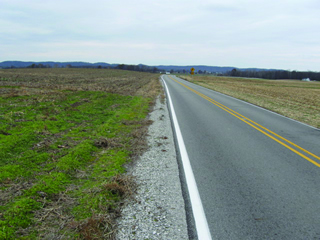 This photograph shows one of the sites that was resurfaced and treated with the safety edge. The road is a narrow, two-lane paved highway with a double yellow line down the middle, a single white line near the edge of the pavement, and fields on both sides of the road.