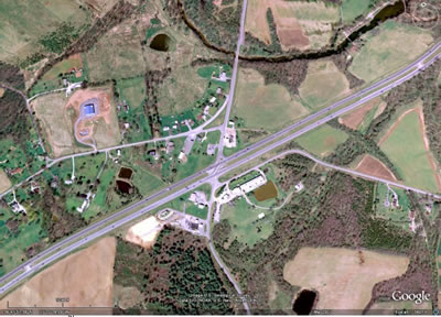 Figure 1. Photo. RCUT intersection observed in this study. This photo shows a satellite view of the restricted crossing U-turn (RCUT) intersection that was the subject of this study. A 
four-lane highway, US-15, is shown extending from the bottom left to the top right. A two-lane rural road, US-15 Business, intersects the highway in the north-south direction. The main intersection has an opening that allows traffic on the divided highway to turn left onto the local road. Curbing can be seen that blocks traffic from the minor road from turning left through the opening at the main intersection. Local road traffic must turn right at the main intersection. Directional U-turn openings can be seen in the highway median 1,800 ft on either side of the main intersection. These openings allow local road traffic to make a U-turn to complete through or left-turn movements.