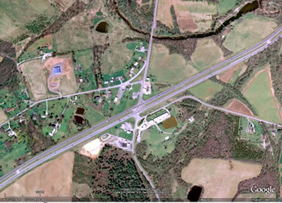 Figure 1. Photo. RCUT intersection observed in this study. This photo shows a satellite view of the restricted crossing U-turn (RCUT) intersection that was the subject of this study. A 
four-lane highway, US-15, is shown extending from the bottom left to the top right. A two-lane rural road, US-15 Business, intersects the highway in the north-south direction. The main intersection has an opening that allows traffic on the divided highway to turn left onto the local road. Curbing can be seen that blocks traffic from the minor road from turning left through the opening at the main intersection. Local road traffic must turn right at the main intersection. Directional U-turn openings can be seen in the highway median 1,800 ft on either side of the main intersection. These openings allow local road traffic to make a U-turn to complete through or left-turn movements.