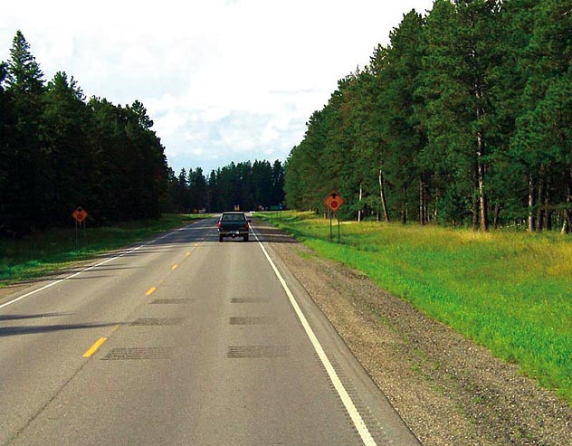 This photo shows a two-lane rural road in Minnesota. There are three sets of transverse rumble strips (TRSs) on the road before a stop ahead warning sign. A motor vehicle is located past the TRSs and is approaching the stop sign.