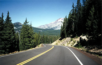 This is a picture of a road running through a mountainous, wooded area. Rocks and trees are shown on either side of the road, and mountains are seen ahead in the distance.