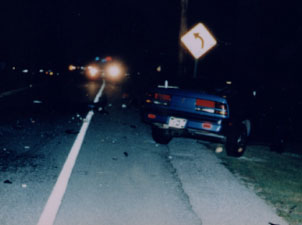 This photograph was taken at night and shows a car that has crashed along the right shoulder of a road. Another vehicle's headlights are shown shining toward the car.