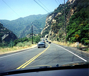 This photograph is taken from the inside of a vehicle that is traveling on a road in the mountains. A mountain is shown along the right side of the road. Several cars are traveling in front of the vehicle from where the photo was taken, and a pickup truck is shown traveling in the opposite direction in the left lane.