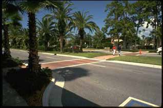 1) Midblock crossing curb extensions provide better visibility for motorists and pedestrians. In this figure, two lanes of traffic are moving in each direction, divided by a landscaped median. There is a bike lane and a lane for parallel parking on the far outsides of the street in both directions. At a midblock crossing point, with a continental-style crosswalk pattern and lit by street lights, there is a curb extension which juts out to the end of the parking lane, not restricting the bike lane. This allows for pedestrians to have a shorter crossing distance across the roadway. 