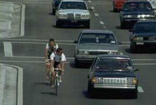 This photograph shows two bicyclists riding single file in a wide curb lane. Vehicles are passing the bicyclists in the same lane but are not crossing the road centerline.