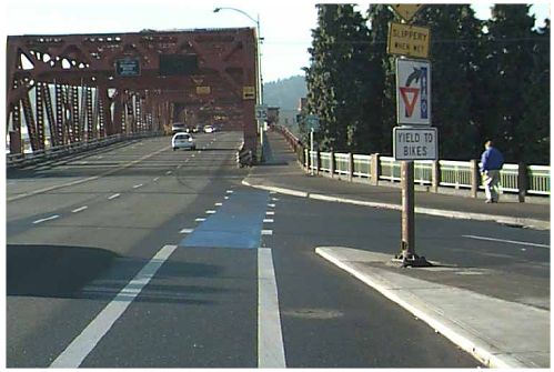 This photograph show the use of a blue bike lane in Portland, Oregon. The pavement surface of the bike lane is colored blue where an entrance ramp crosses it. A sign on the entrance ramp indicates that motor vehicles are to yield to bikes in the blue bike lane.