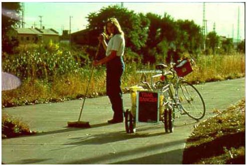 This photograph shows a woman with a push broom who is cleaning a wide shared use path. Her bicycle is also parked on the path and include a trailer that contains maintenance equipment.
