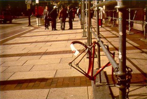 This photograph shows the frame of a bicycle that has been locked to a railing made of steel pipe. Apparently the wheels, seat, and most of the bike parts and accessories has been stolen while the bike was parked at this location.