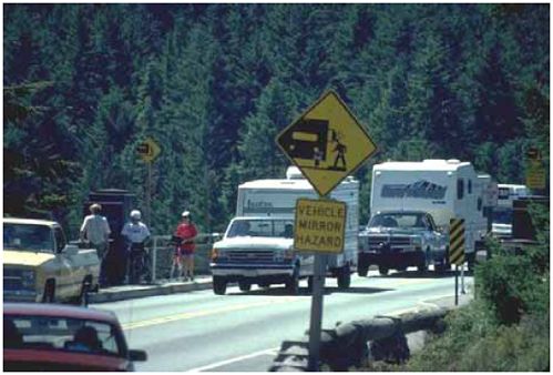 The photograph shows busy roadway with several large trucks towing recreational campers. There is a sidewalk on both sides of the road when crossing a bridge, but the sidewalks have not been given adequate clearance from the travel lanes. A warning sign at the beginning of the bridge shows a symbol of a vehicle mirror striking a pedestrian with the text - VEHICLE MIRROR HAZARD.