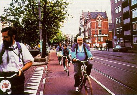 This photograph shows a wide bicycle lane that has a red pavement color. Three bicyclists are shown riding single file in the lane, with another dismounted bicyclist in the foreground of the photo.