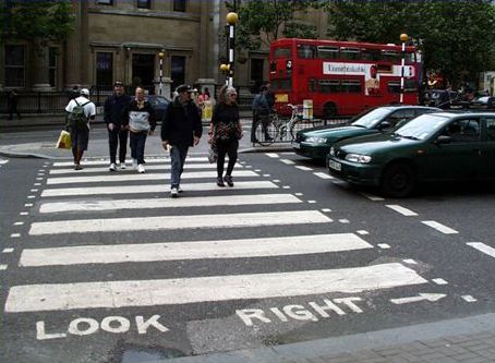 The photograph shows a marked pedestrian crosswalk with a text message 'LOOK RIGHT' painted on the pavement surface right next to the curb. Several people are crossing in the crosswalk and two vehicles are waiting in back of the crosswalk.