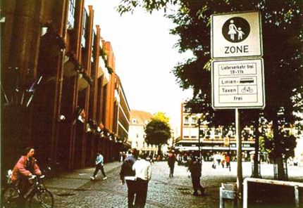 This photograph shows a street in Germany that operates as a pedestrian mall during daytime areas. Many pedestrians can be seen crossing or walking in the street and a bicyclist is crossing the street.