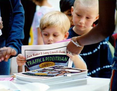 The 5 E’s. The photograph shows a young child being handed traffic safety education materials in a classroom setting.