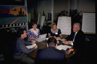 The photograph shows five adults sitting around a table and discussing issues. All people are intently listening to the person speaking.