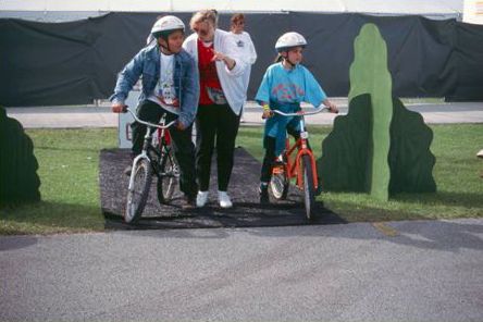 The photograph shows two young children on bikes being instructed by a standing adult. The adult and children appear to be standing in a bike safety course.