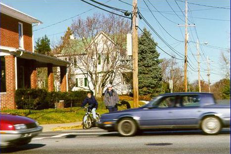 In this photo, a young child on a bike is waiting with his father on a curb in a neighborhood. Both are looking at the cars going in both directions on the street in front of them.