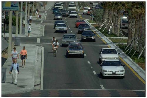 Photo shows bicyclist riding without helmets. They are biking along side traffic on a 2 lane road.