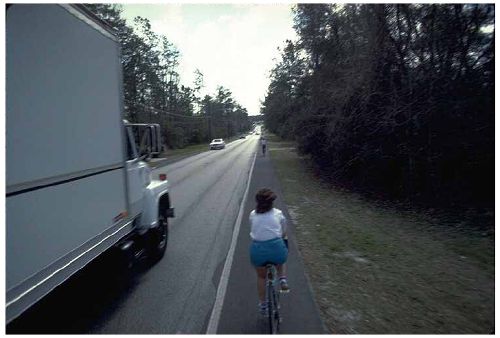 This picture shows a bicyclist not wearing a helmets while biking on the side of road with a truck passing by.