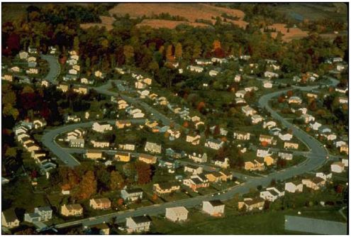 There is an aerial photograph of a suburban development. The streets curve and snake through the landscape, and by design, they do not lend themselves to easy pedestrian movement. 