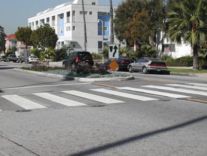 This photo shows a view from the sky of a zebra crossing (two parallel lines running perpendicular to the roadway with diagonal lines inside the outer two) with in-roadway warning lights located on the outside of the crosswalk.