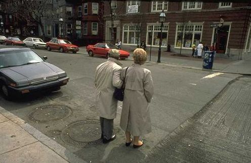 Reduced visibility of pedestrians behind parked cars can create conflict. In this photo, an older man and woman stand at the front of a car parked on the side of the road and look to their left to scan for any cars coming from behind the row of parked cars.