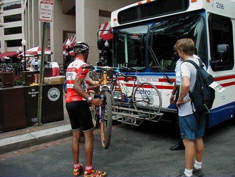 Picture shows two men loading their bicycles onto the front bicycle rack of a city bus.