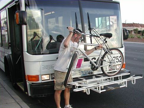 Picture shows a man loading a bicycle onto a front rack of a city bus.