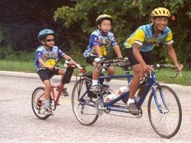 Second picture shows a father and child on a tandem bike pulling a second child in a trail-a-bike.