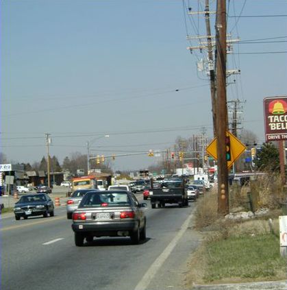 Picture shows cars traveling on a busy street through a strip-style commercial area. There are no pedestrians, no sidewalks and no shoulder lanes to walk on.