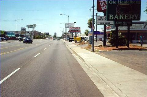 Picture shows a broad, 5-lane road in strip shopping center. There is a sidewalk between the parking lot driveways and street. There are no trees or any landscaping. There are very few cars, and the road seems over-built.