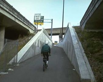 The picture shows a bicyclist entering a walled section of a shared use trail. Numerous trail underpass and bridges are present in the picture.