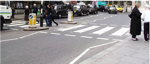 Zebra crossing in London with zigzag approach markings and Belisha beacons. This photograph shows a pedestrian crossing with continental-style pavement markings, a median refuge island, and small posts with a globe that glows when activated by pedestrians (belisha beacon).