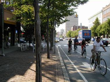 The picture shows a tree-lined downtown street with a bike lane and four bicyclists riding in the bike lane in the direction of the camera.