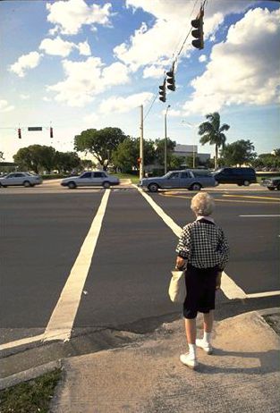 The picture is of an elderly woman standing at the curb, waiting to cross the street. She is standing at the end of a very long crosswalk with no crossing refuge in the middle.