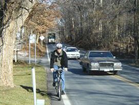 The  picture is of a person riding a bicycle on the narrow paved shoulder of a busy country road.