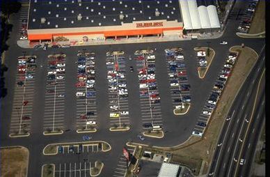 The picture on the left is an aerial view of a parking lot of a shopping center. The parking lot is half filled with cars. In order to get to the shopping area, someone must park and walk through the parking area.