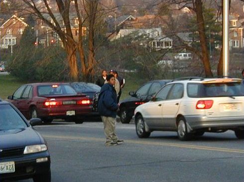 The picture shows a photo of a boy, standing on the yellow line in the middle of a road, attempting to make a mid block crossing in busy two-way traffic. 